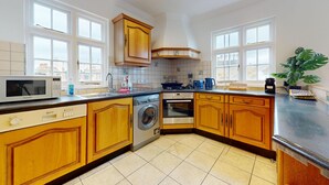 A kitchen with wooden cabinets, a washing machine, a microwave, and a coffee maker. - Bright 2BR Apt- 11 min walk to Kensington Palace (London)