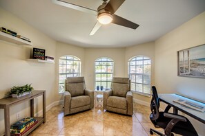 Una habitación con ventilador de techo, dos sillones, una mesita y un escritorio con silla. Se ven libros en una estantería y una planta sobre una mesa.