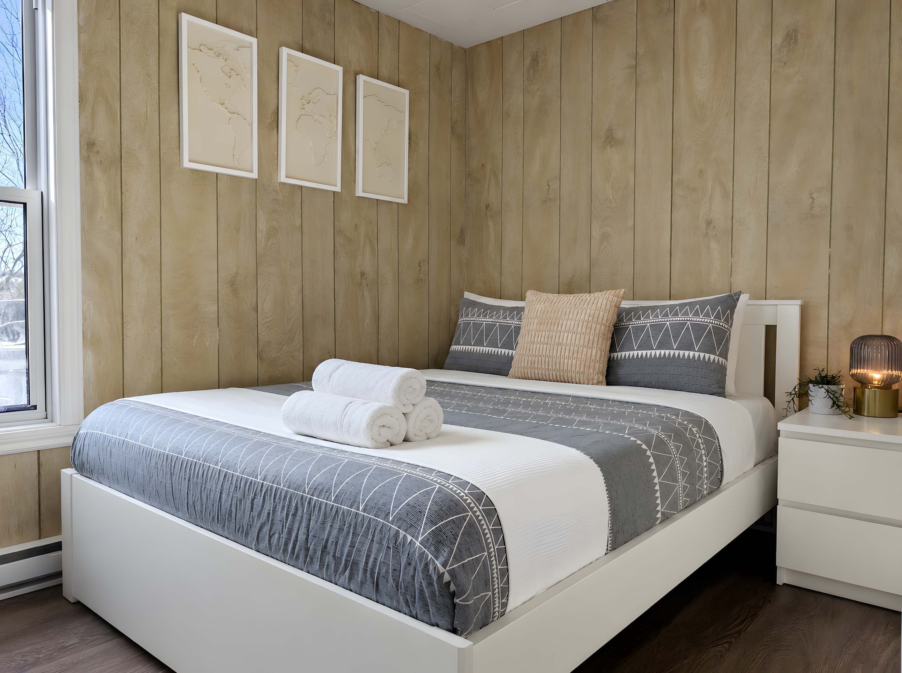 A bedroom with a wooden wall, a bed with a patterned bedspread, two pillows, a bedside table with a lamp, and three framed pictures on the wall.