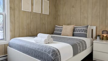 A bedroom with a wooden wall, a bed with a patterned bedspread, two pillows, a bedside table with a lamp, and three framed pictures on the wall.