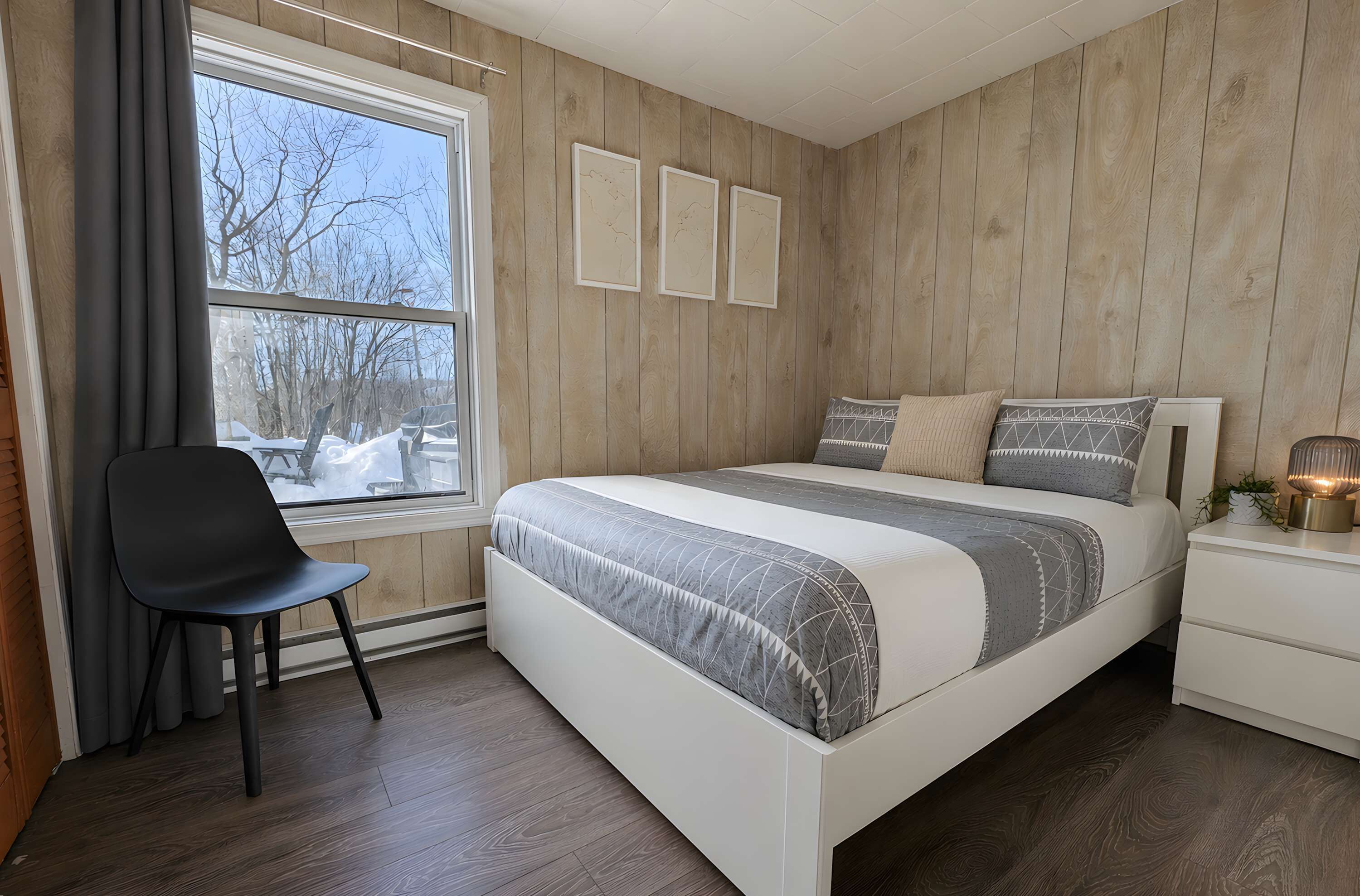 Een slaapkamer met een bed, een stoel, een nachtkastje, een raam met gordijnen en houten lambrisering.