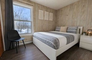 Ein Schlafzimmer mit einem Bett, einem Sessel, einem Nachttisch, einem Fenster mit Vorhängen und Holzwandverkleidung.