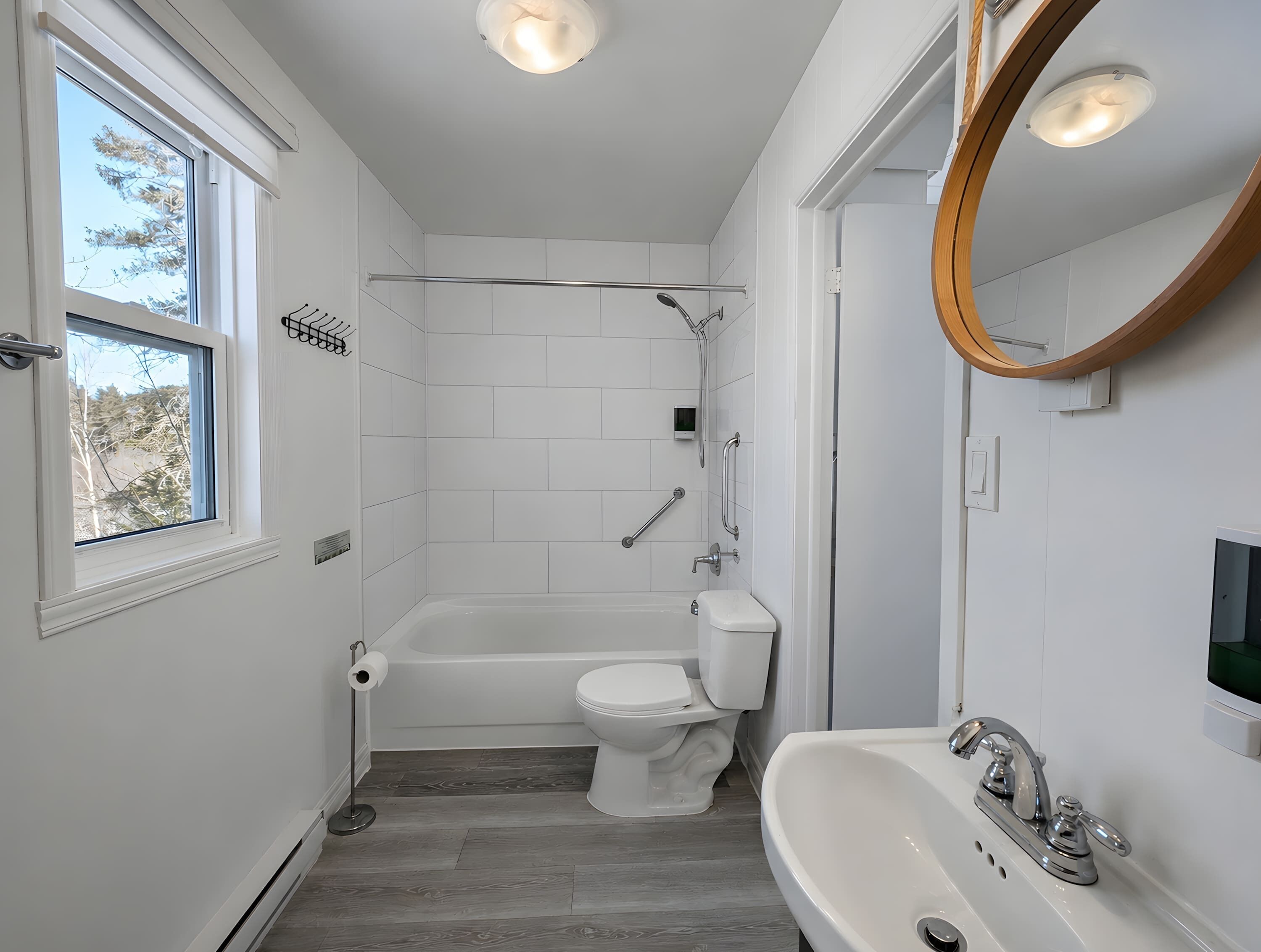 Une salle de bain moderne équipée d’une baignoire, de toilettes et d’un lavabo, avec des carreaux blancs et une grande fenêtre.