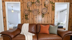 Un canapé en cuir marron, agrémenté d’un plaid blanc et de deux coussins, est placé dans une pièce aux murs recouverts de lambris en bois. On y aperçoit également un luminaire suspendu et, au fond, un lit avec une carte accrochée au mur.