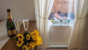 Una botella de champán, dos copas y un ramo de girasoles sobre una mesa junto a una ventana con cortinas.