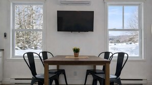 Un coin repas comprenant une table, quatre chaises, un téléviseur à écran plat fixé au mur et de grandes fenêtres donnant sur un paysage enneigé.