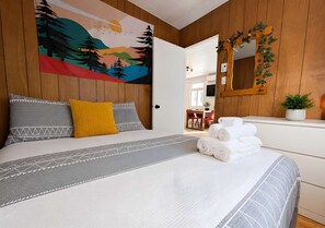 A bedroom with a bed, pillows, a blanket, a wall tapestry, a mirror, and a plant. - Le Grand Pin | 6 pers | Spa | Massif & MSA | 16 (Saint-Tite-des-Caps)