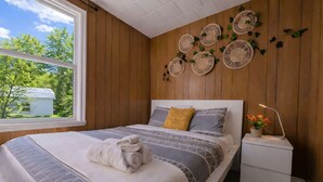 A bedroom with a large window, wooden walls, a bed with patterned bedding, decorative wall hangings, and a bedside table with a lamp.