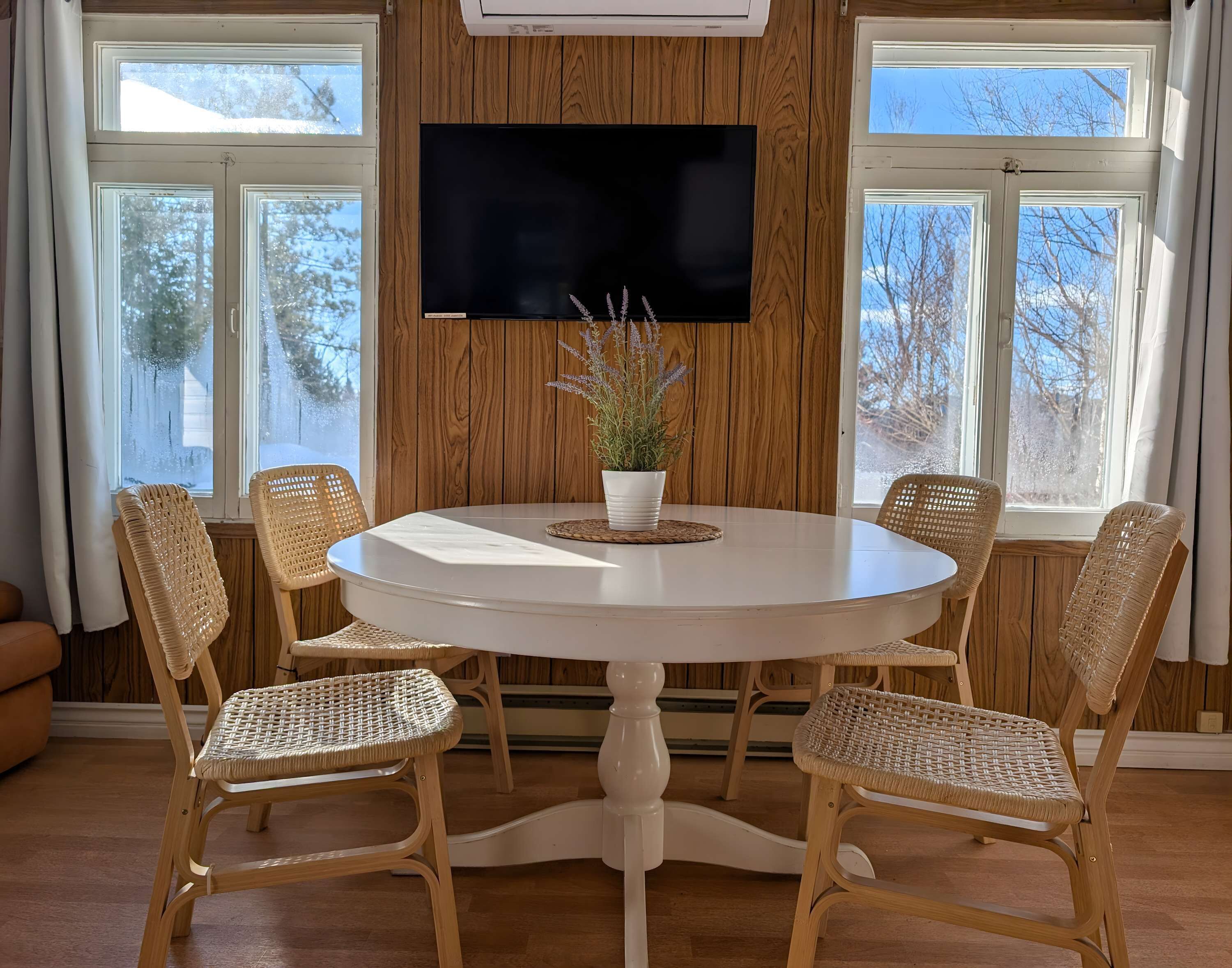 A round dining table with a white tablecloth, surrounded by four wicker chairs, a potted plant, and a flat-screen TV mounted on a wooden wall.