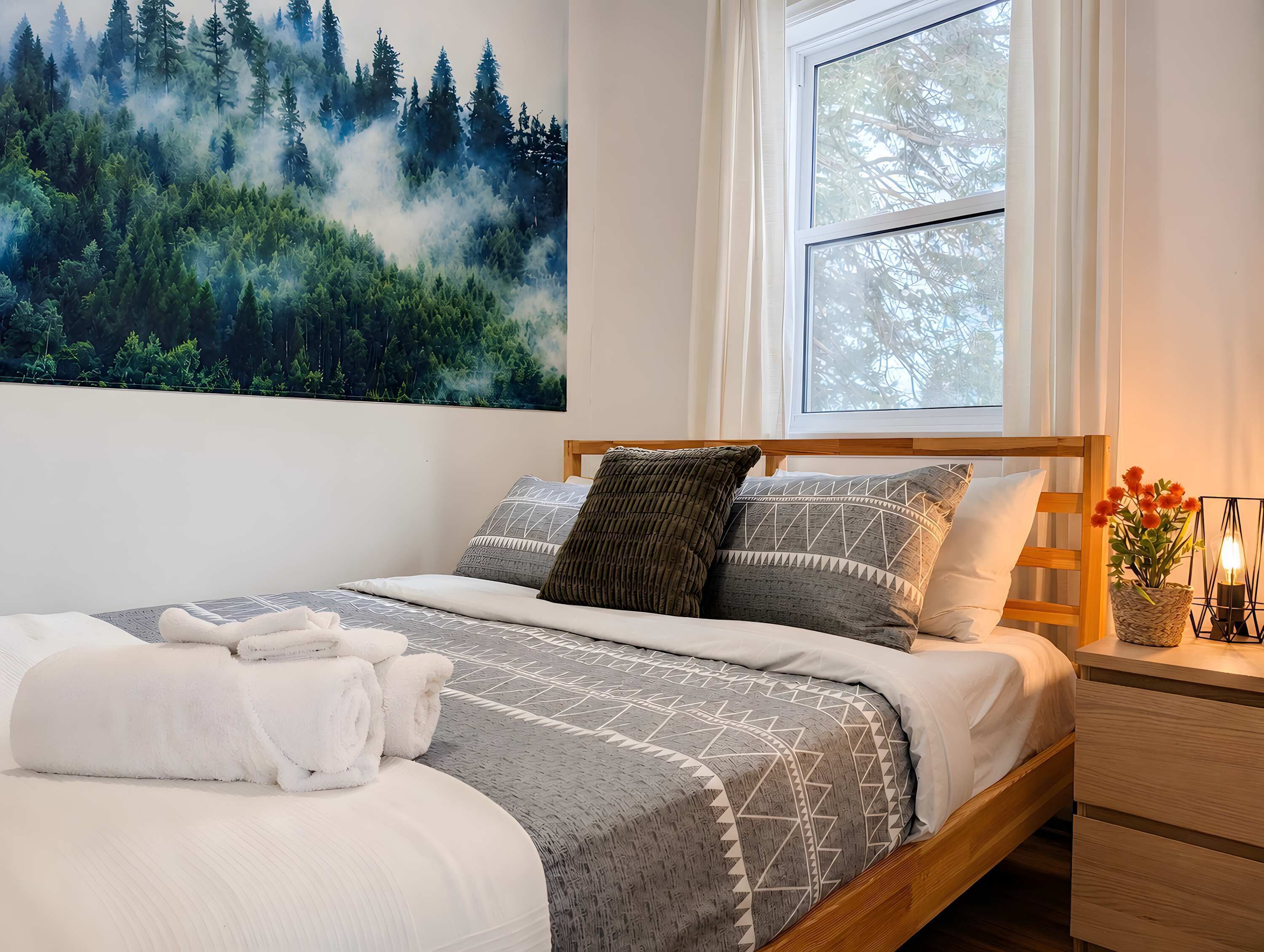 Un lit bien fait, recouvert d’une couvre-lit à motifs, une fenêtre avec des rideaux, un tableau représentant une forêt accroché au mur, et une table de chevet ornée d’un vase de fleurs.