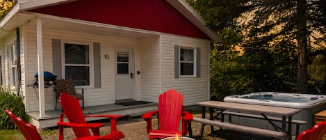 Una cabaña pequeña con un porche, sillas Adirondack rojas, un jacuzzi y una fogata.