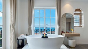 A modern bathroom with a large freestanding bathtub, a vanity with a mirror, and a view of the sea through a large window. - Amaris Hotel (Phu Quoc)