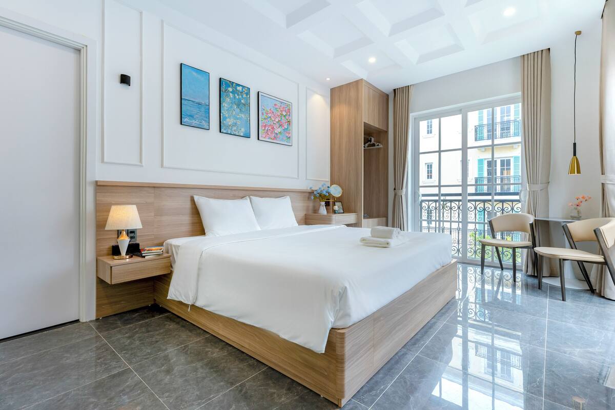 Habitación de hotel con una cama grande, una mesita de noche con lámpara, una pequeña mesa con sillas y un balcón con vistas a edificios.