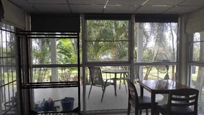 Een kamer met een tafel en stoelen, een bank met spullen erop en grote ramen met uitzicht op groen.