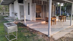 Een overdekt buitengebied met een barbecue, een tafel met stoelen en een wit gebouw met een deur en raam.