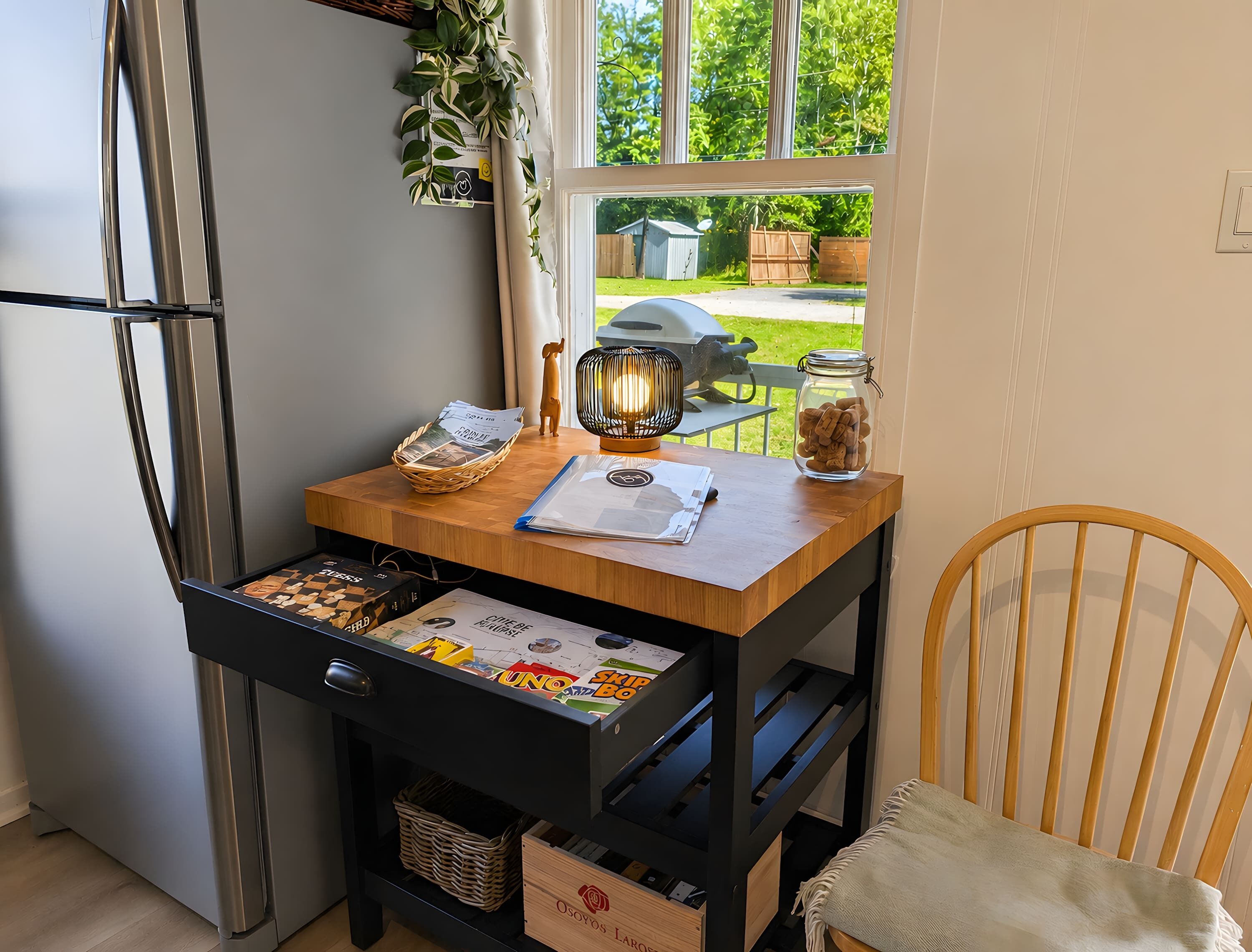 A kitchen with a wooden table, a refrigerator, a wicker basket, a jar of cookies, and a window with a view of the outdoors.