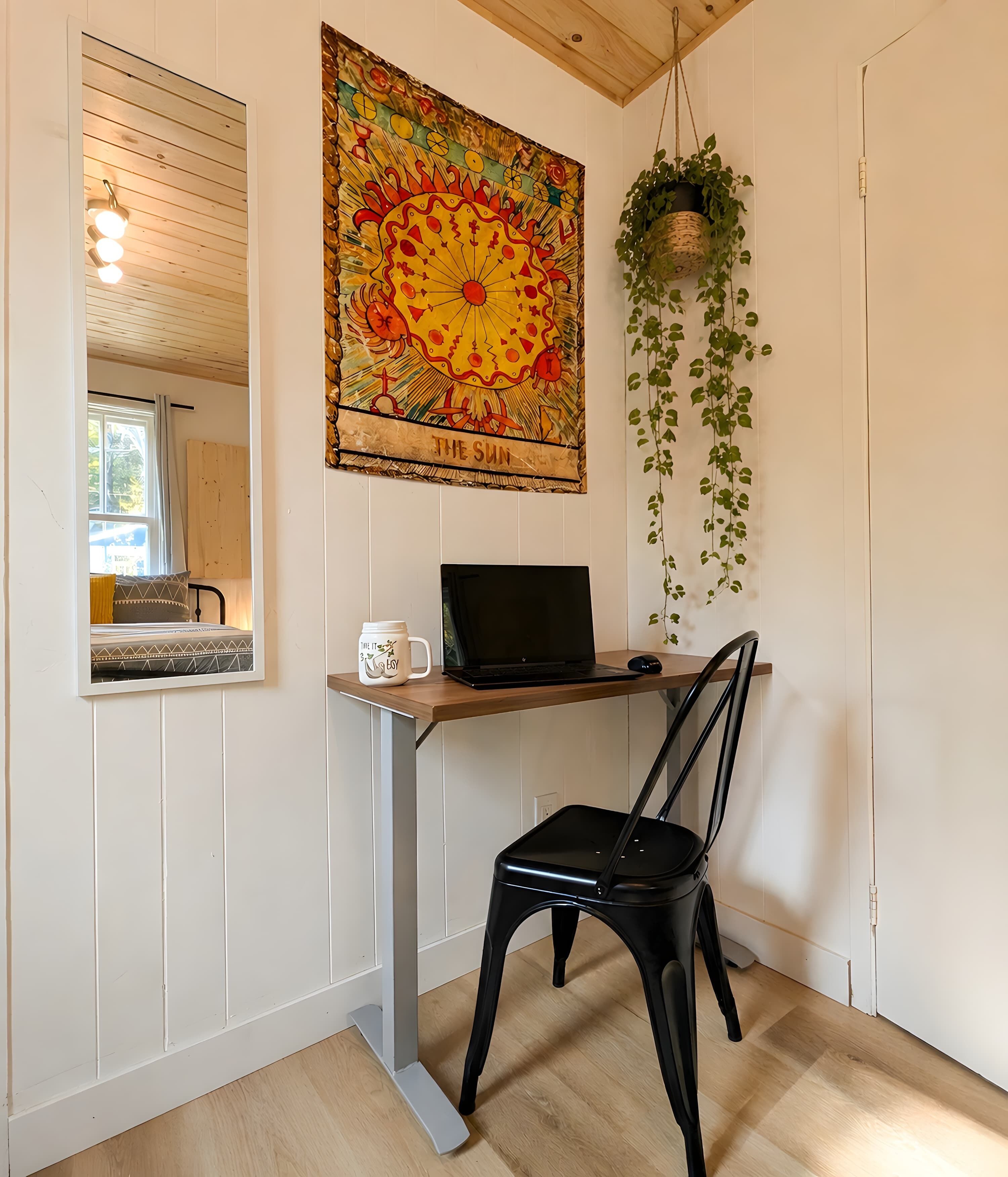 A room with a wooden desk, a laptop, a black chair, a window with blinds, and a wall-mounted painting.