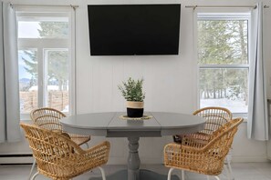 Ein Essbereich mit einem runden Tisch, vier Korbstühlen, einer Topfpflanze und einem großen Flachbildfernseher an der Wand.