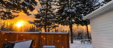 Un jacuzzi en un patio nevado con valla de madera, árboles y una casa.