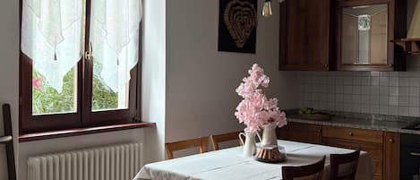 A dining area with a table set for four, a radiator, a window with curtains, and a kitchen in the background.