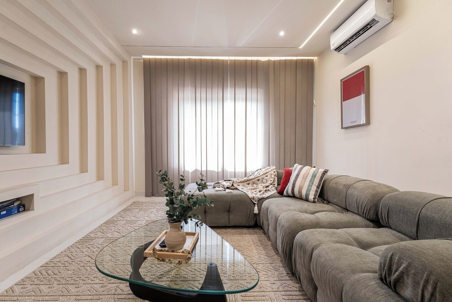 Una sala moderna con un sofá seccional, una mesa de centro de vidrio y un televisor montado en la pared.