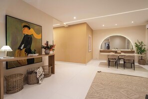 Una sala moderna con un cuadro grande, una consola con adornos, cestas de mimbre, una zona de comedor con sillas y un espejo grande.