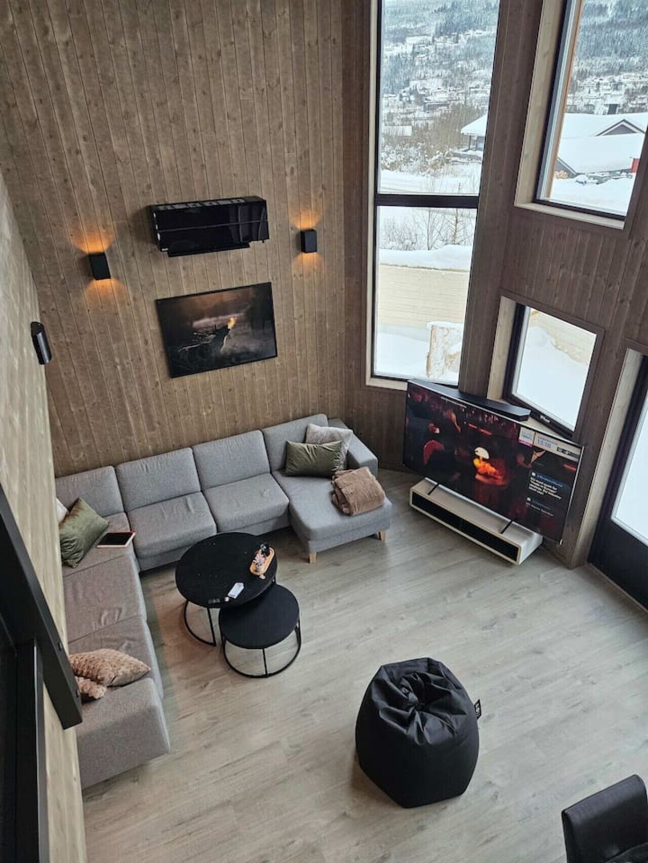 A modern living room with a grey sectional sofa, a black coffee table, a black bean bag, a flat-screen TV, and a wooden wall with built-in speakers.