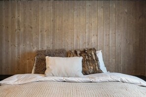 Ein Bett mit weißer Bettwäsche und zwei Kissen, eines davon mit fellartiger Struktur, vor einer Wand mit Holzverkleidung.