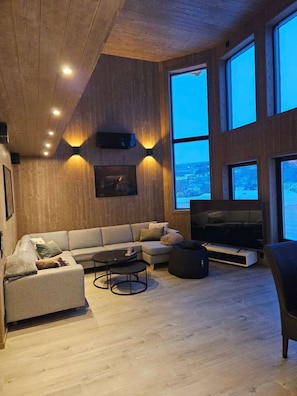 A modern living room with a sofa, a coffee table, a black bean bag, a flat-screen TV, and large windows offering a view of the outdoors.