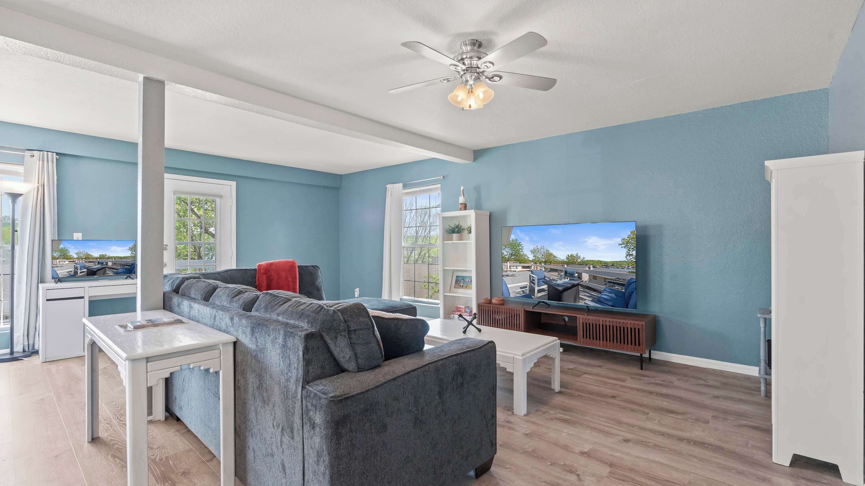 A spacious living room with a grey sectional sofa, a white coffee table, a ceiling fan, and a large flat-screen TV mounted on the wall.