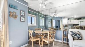 A dining area with a round wooden table and chairs, a ceiling fan, and a kitchen area visible through a glass partition. - Gulf Shores Plantation, 5508 - Beachfront Condo with Gulf Views (Gulf Shores)