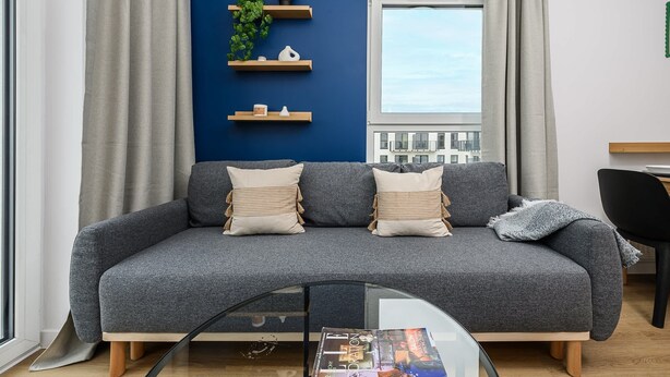 Una sala de estar moderna con un sofá gris, dos almohadas, una mesa de centro de vidrio y una ventana con cortinas.
