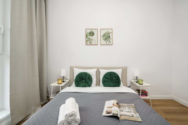 A neatly made bed with a grey quilt, two green decorative pillows, a white headboard, and two framed botanical prints on the wall.