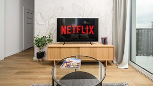 Una sala de estar moderna con una televisión mostrando 'NETFLIX', un mueble de madera, una mesa de centro de vidrio con una revista y una planta.