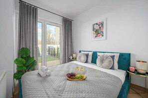 Un dormitorio con una cama, una ventana con cortinas, una planta, una mesita de noche con una lámpara y un cuadro en la pared.