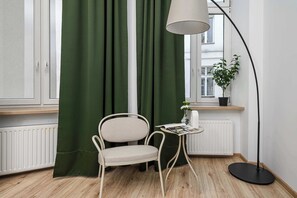 Ein Zimmer mit einem Sessel, einem Tisch mit einer Zeitschrift, einer Stehlampe, einem Fenster mit Ausblick und einer Heizung.