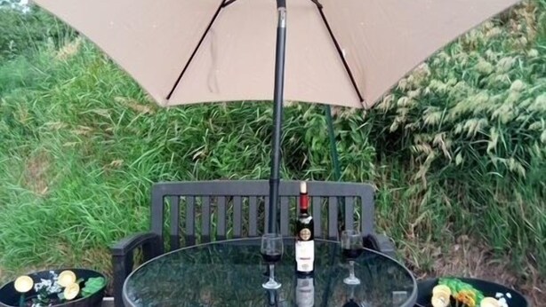 Een ronde glazen tafel met metalen frame, een fles wijn en twee glazen erop, onder een beige parasol.