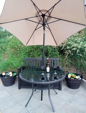 A round glass table with a metal frame, a single bottle of wine, and two glasses on it, under a beige umbrella.