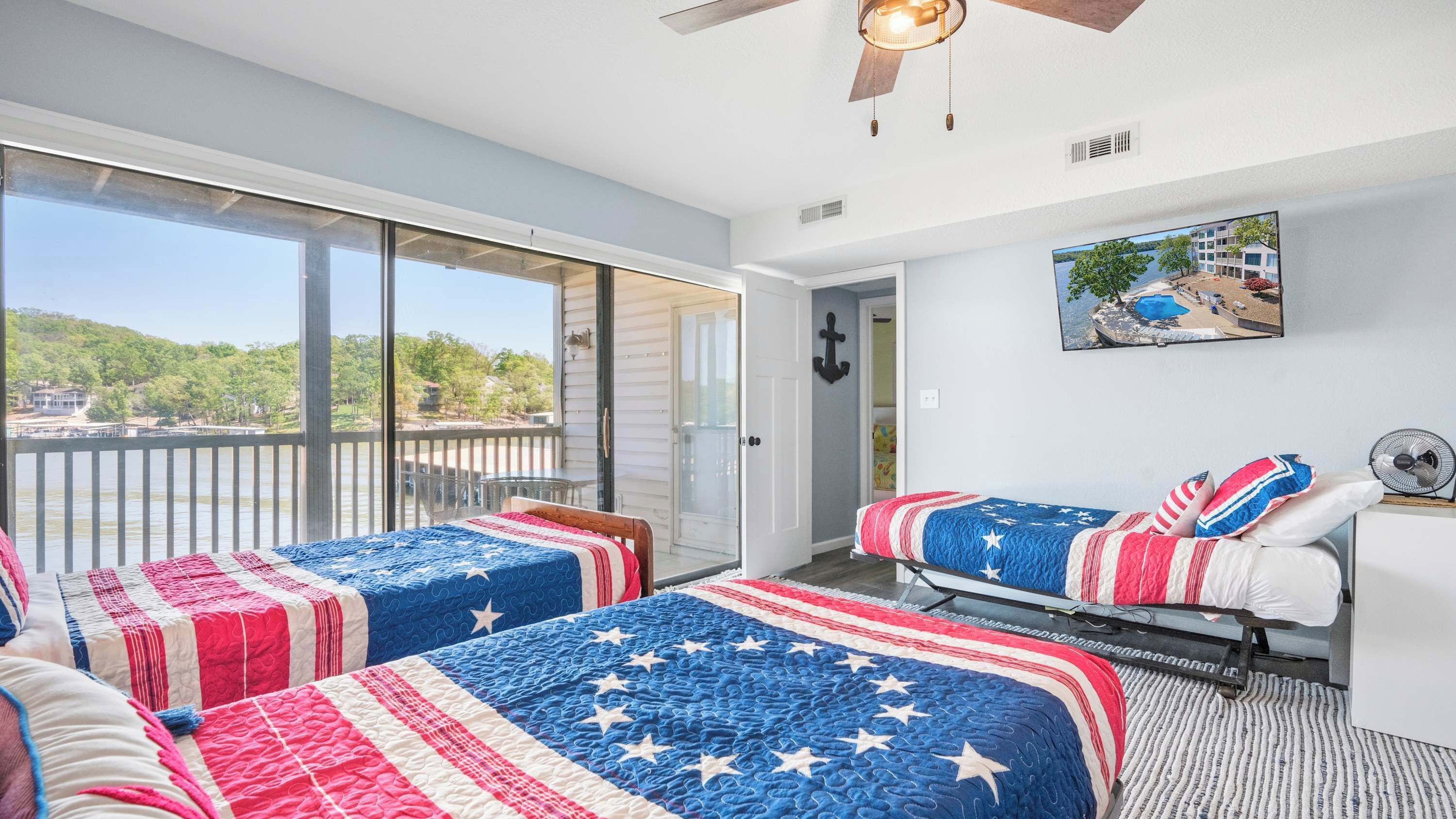 A room with two beds, a balcony with a view of a lake, a ceiling fan, and a painting on the wall.