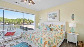A bedroom with a large window, a bed with colorful bedding, a chair with a pillow, a nightstand, and a lamp.