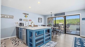 A kitchen with a blue island, a dining area with chairs, a large sliding glass door to a deck, and various wall decorations. - Reel Relaxation Waterfront (Lake Ozark)
