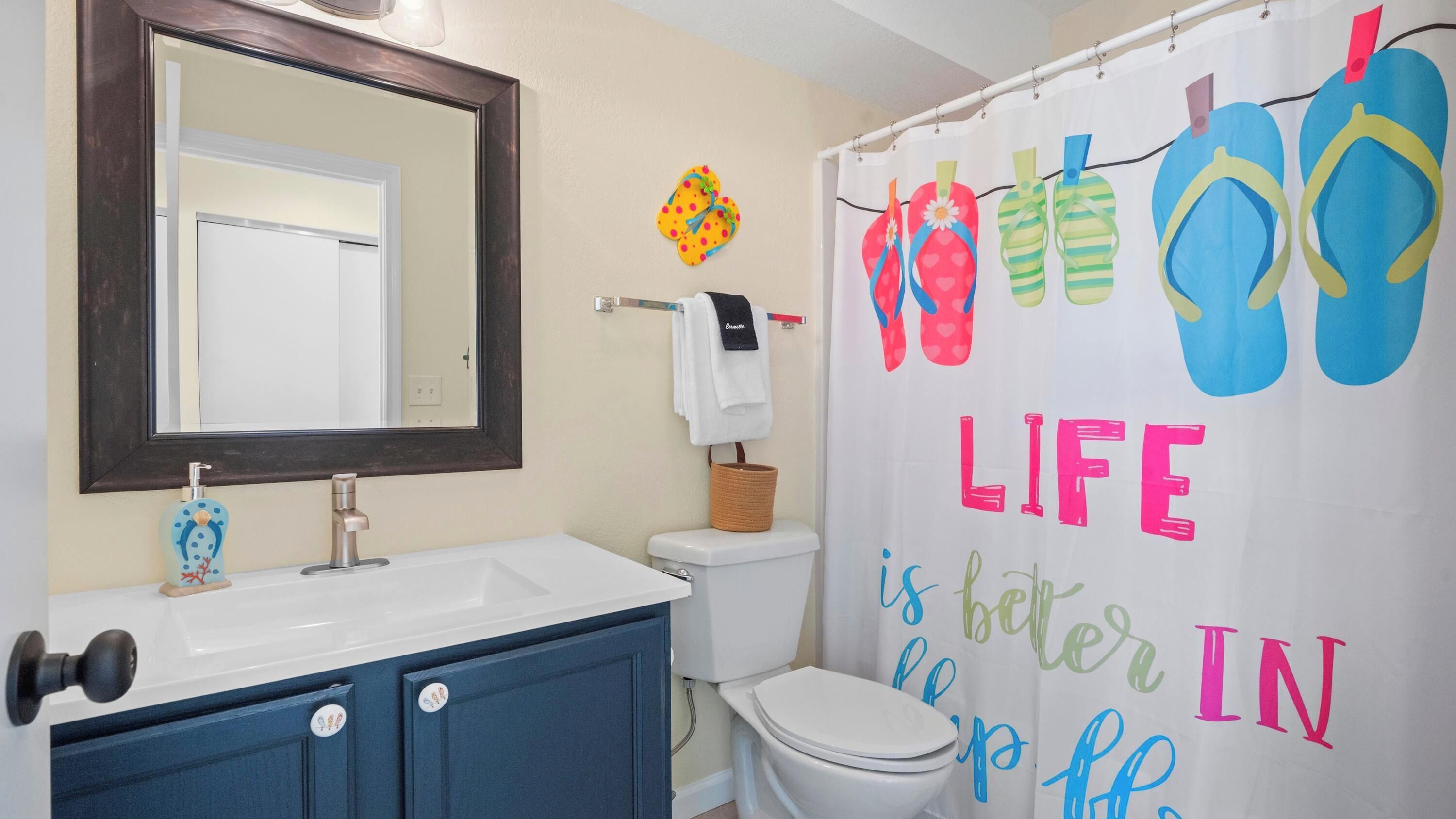 A bathroom with a white sink, a mirror, a toilet, and a shower curtain with flip-flops and the phrase 'LIFE is better in the sun.'