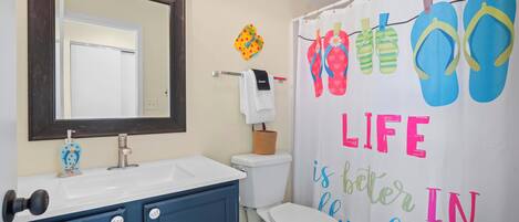 A bathroom with a white sink, a mirror, a toilet, and a shower curtain with flip-flops and the phrase 'LIFE is better in the sun.'