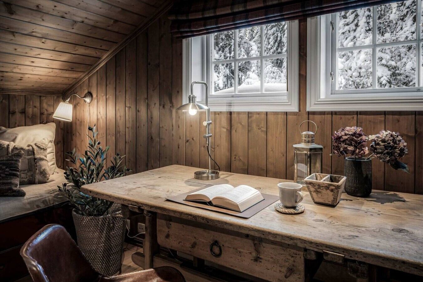 A cozy cabin interior with a wooden desk, a lamp, a book, a cup, and a vase with dried flowers.