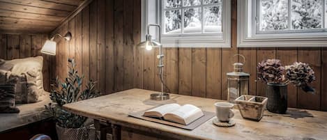 A cozy cabin interior with a wooden desk, a lamp, a book, a cup, and a vase with dried flowers.