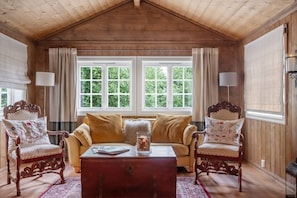 A cozy room with a wooden ceiling, a sofa, two armchairs, a table, and a window with curtains.