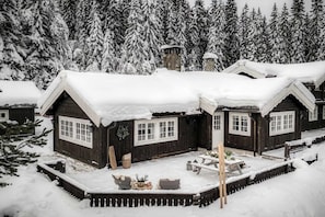 A wooden cabin with a snow-covered roof, large windows, and a small porch area with outdoor seating.