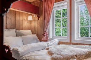 A bedroom with a wooden ceiling, a bed with white bedding, a window with a view of greenery, and a lamp on the wall.