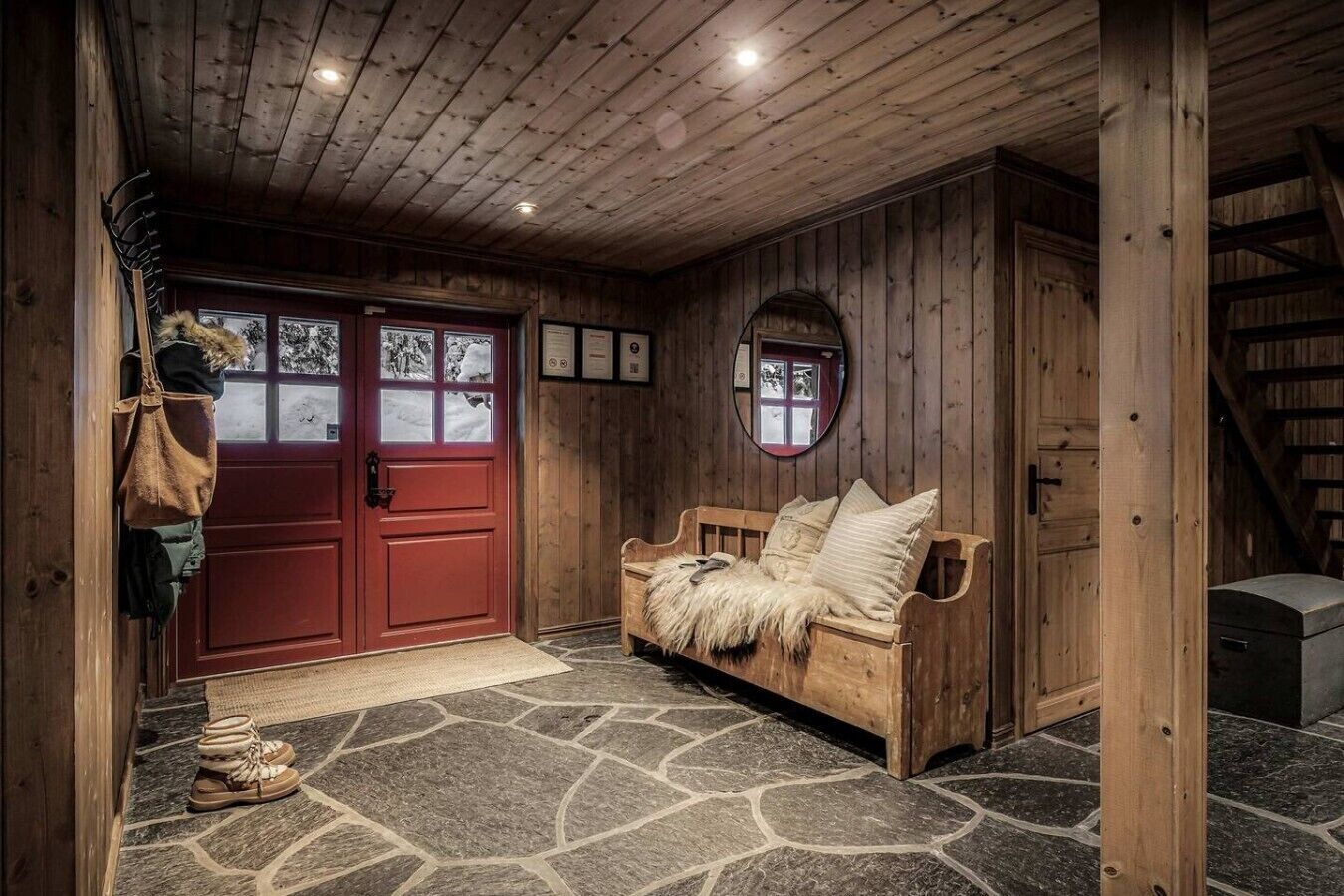 Un intérieur en bois avec une porte rouge, un sol en pierre, un banc en bois et un miroir accroché au mur.