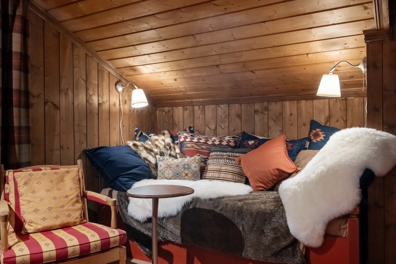 A cozy cabin interior with a wooden ceiling, a sofa with patterned cushions, a chair, and a small round table.